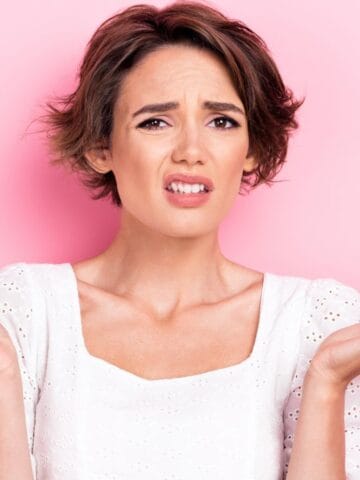 A person with short brown hair wearing a white blouse stands in front of a pink background, holding both hands up and looking confused or uncertain.
