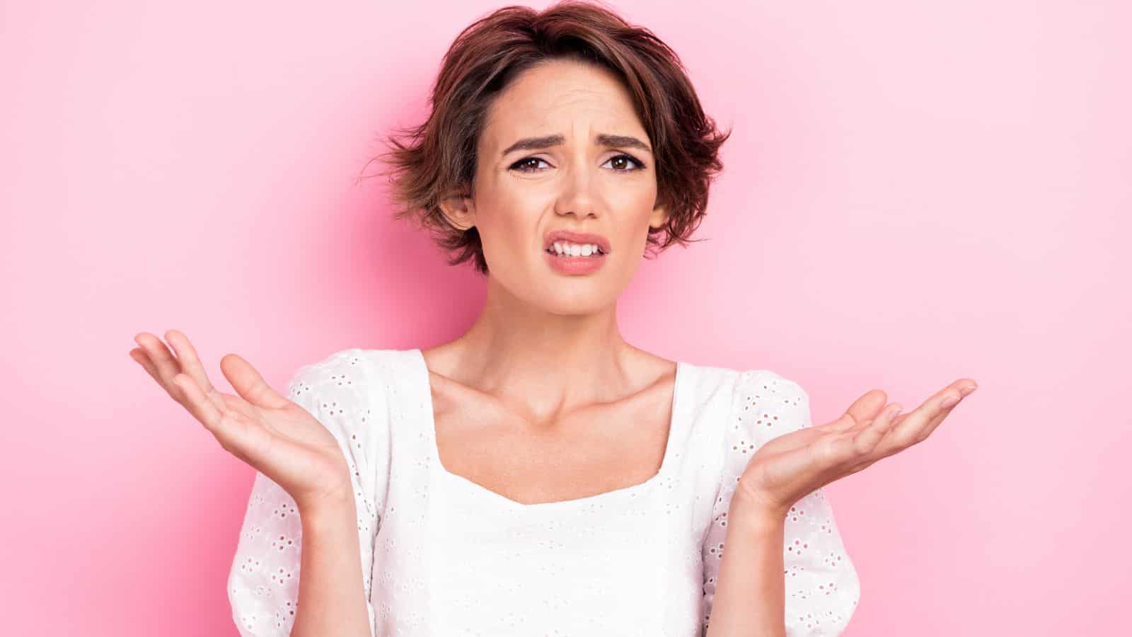 A person with short brown hair wearing a white blouse stands in front of a pink background, holding both hands up and looking confused or uncertain.