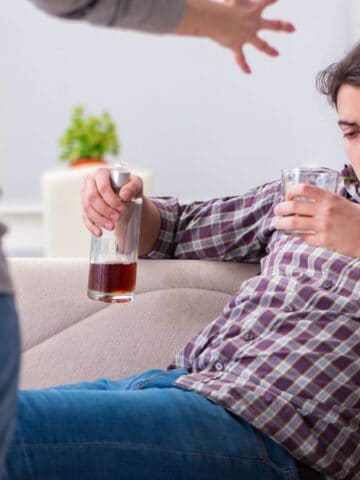A man sits on a sofa holding a glass and a bottle of alcohol, looking down. Another person stands nearby with an arm raised, gesturing toward him in what appears to be a confrontational manner.