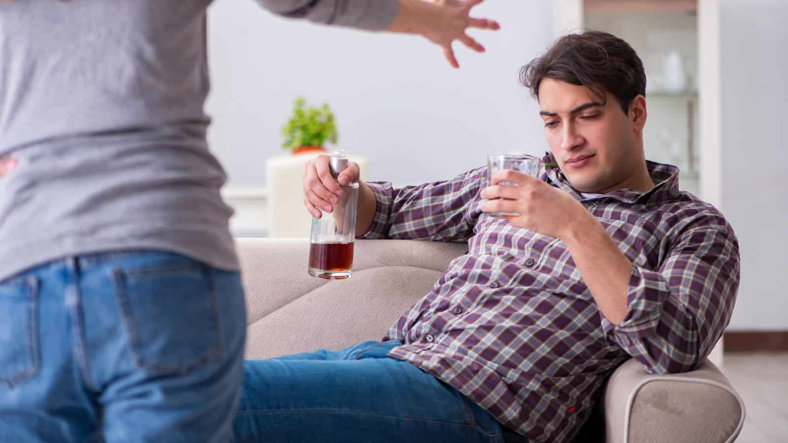 A man sits on a sofa holding a glass and a bottle of alcohol, looking down. Another person stands nearby with an arm raised, gesturing toward him in what appears to be a confrontational manner.