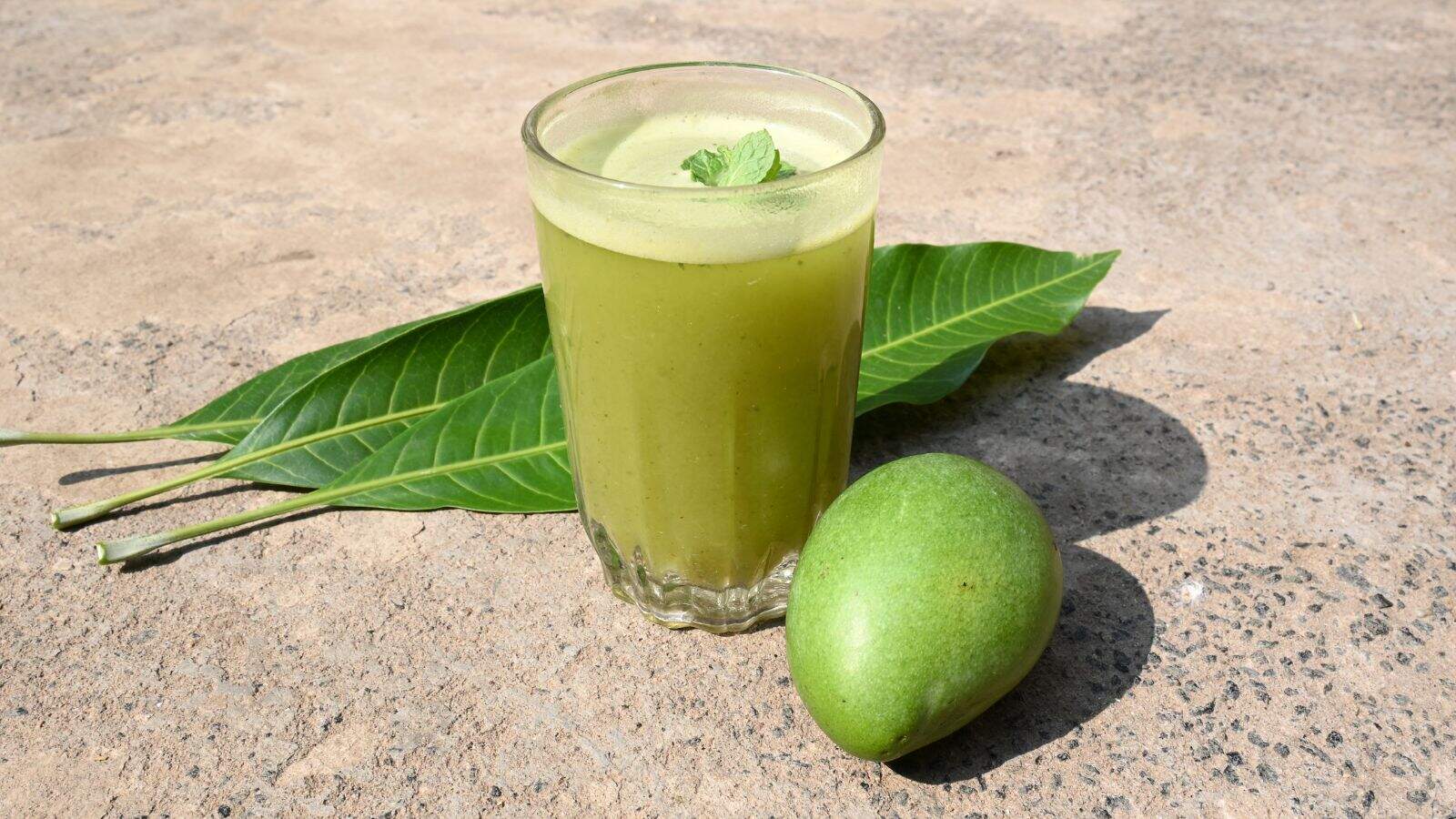 A glass of green mango juice with a mint leaf on top is placed on a flat surface, next to two green leaves and a whole raw green mango.