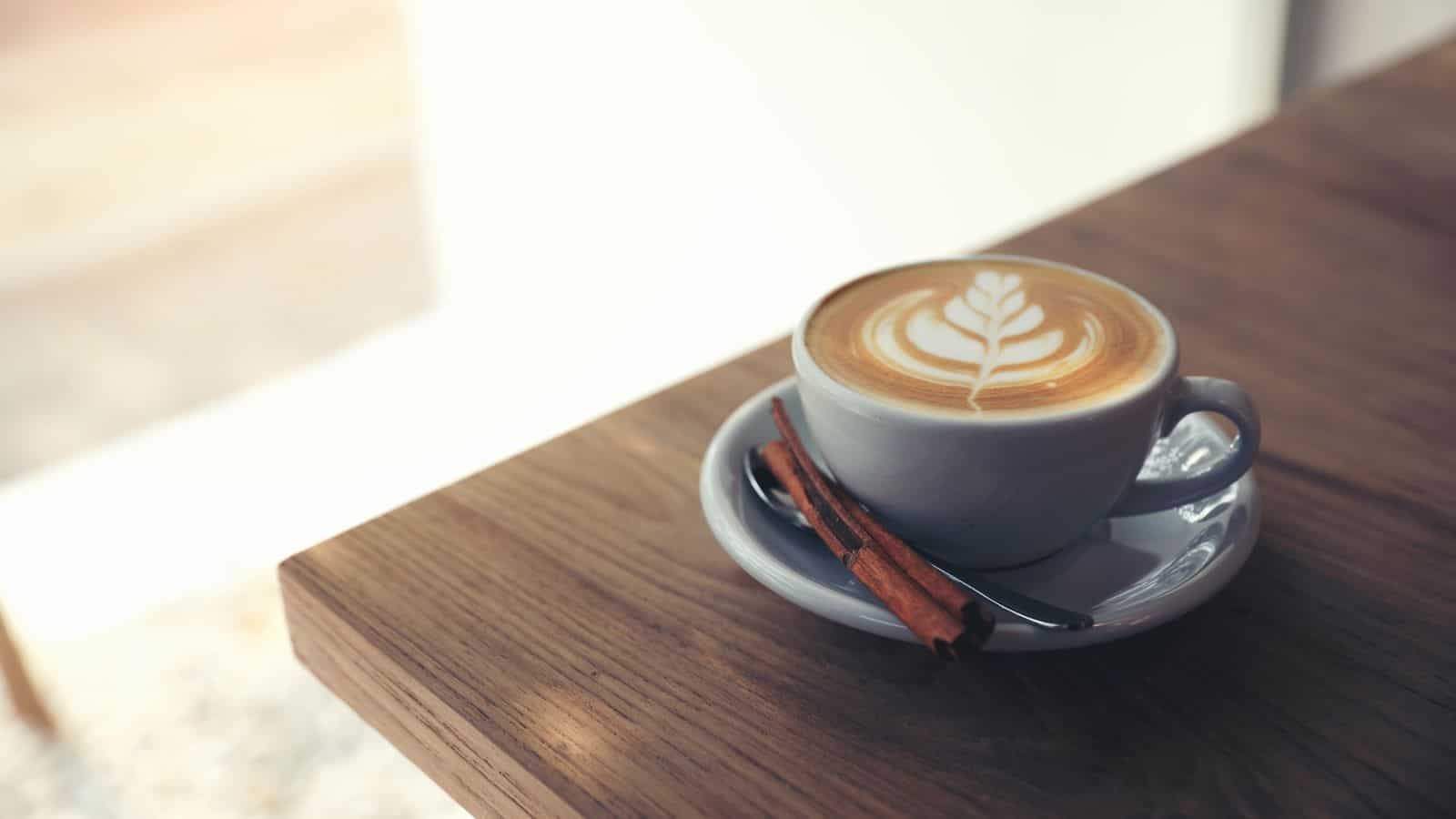 A cup of cappuccino with latte art sits on a saucer beside a cinnamon stick and a spoon on a wooden table, with sunlight coming from the background.