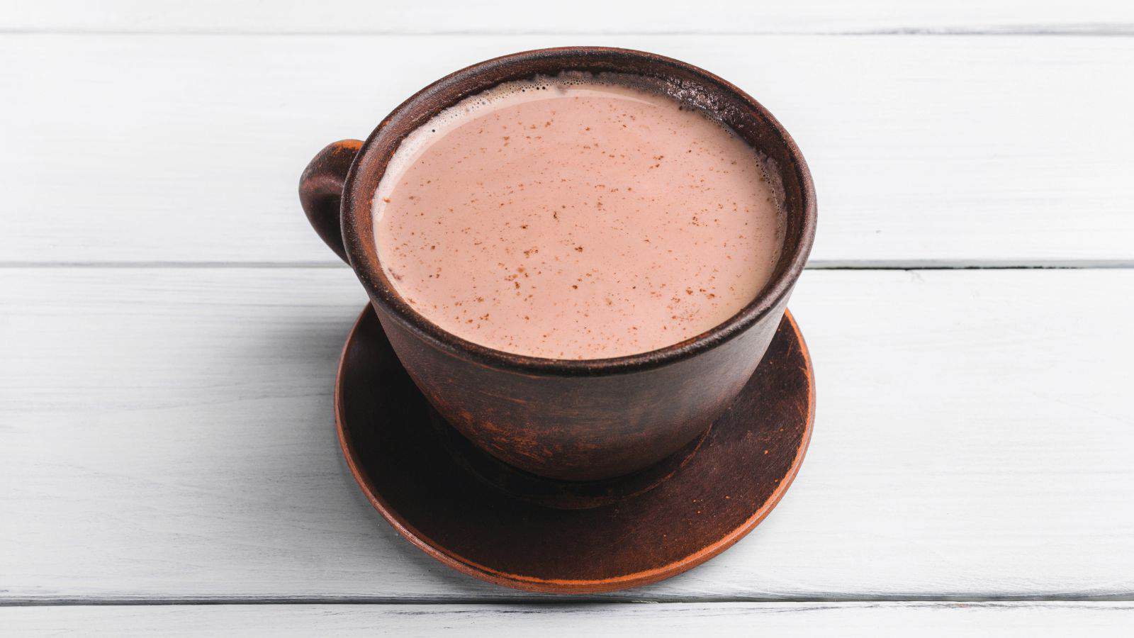 A brown ceramic cup filled with light brown hot chocolate sits on a matching saucer on a white wooden surface.