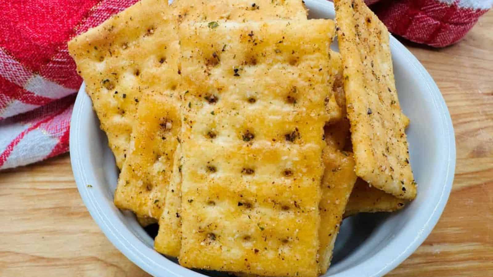 A white bowl filled with seasoned square crackers is placed on a wooden surface, with a red and white checkered cloth in the background.