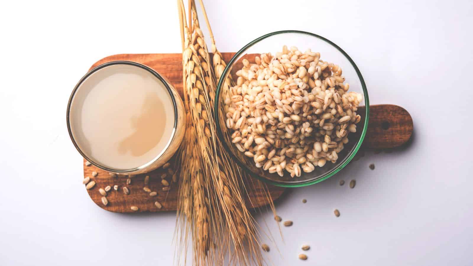 A glass of barley water, a bowl of cooked barley grains, and dry barley stalks are arranged on a wooden board against a white background.