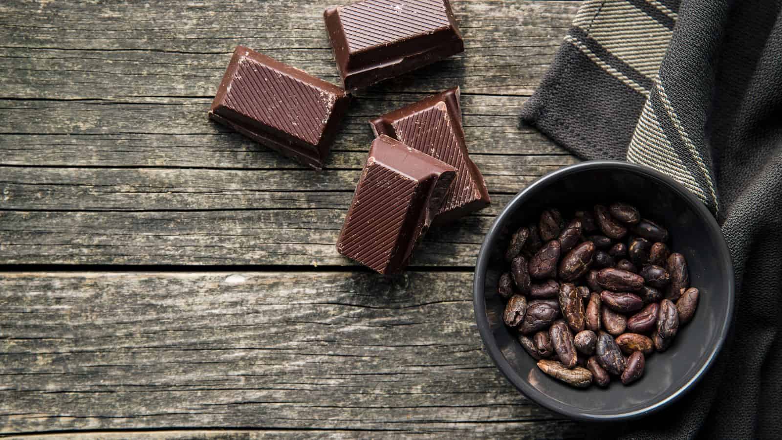 A bowl of cocoa beans next to several pieces of dark chocolate on a rustic wooden surface with a striped cloth nearby.