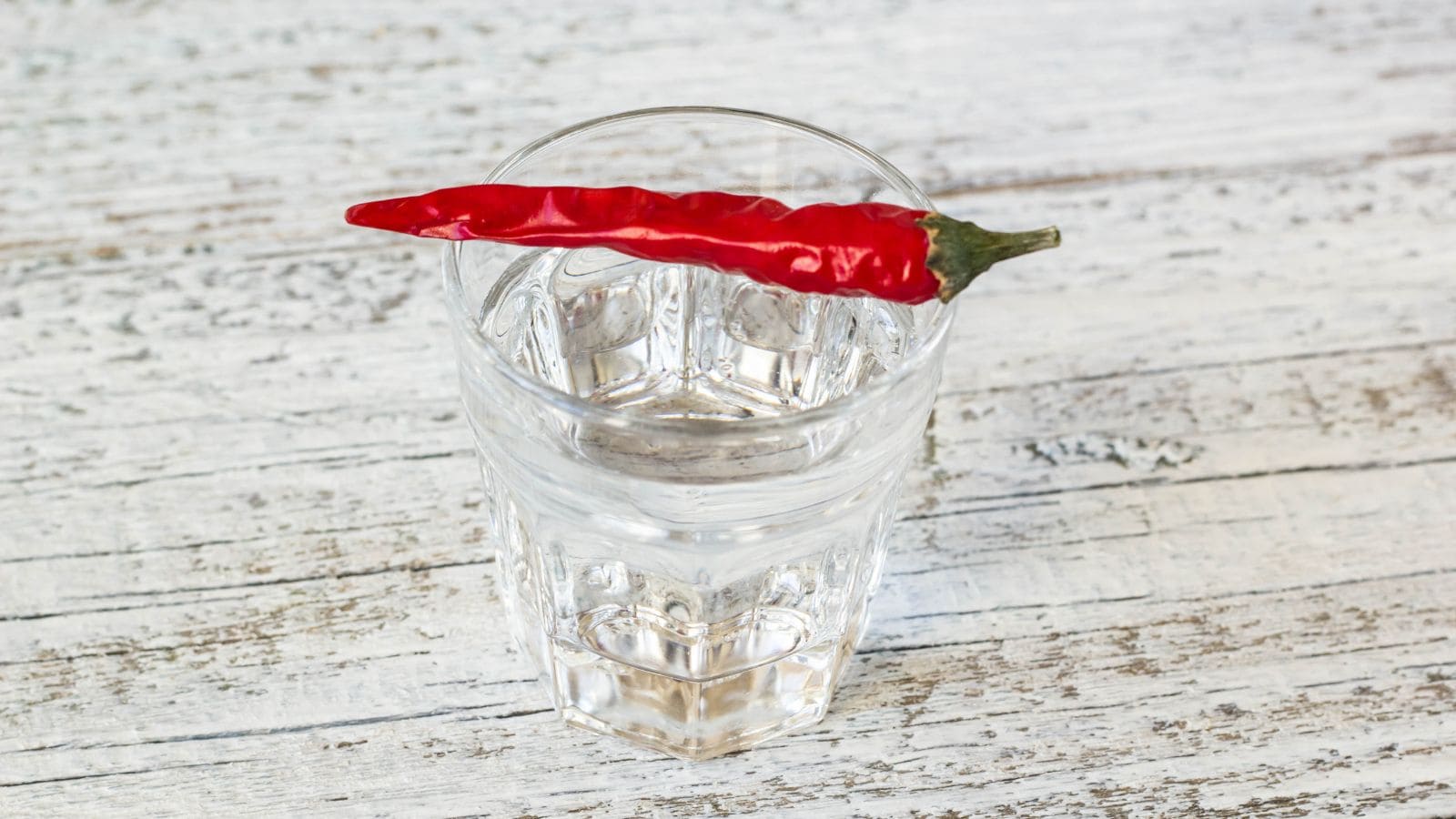 A clear glass of water sits on a white wooden surface with a single red chili pepper resting across the rim of the glass.
