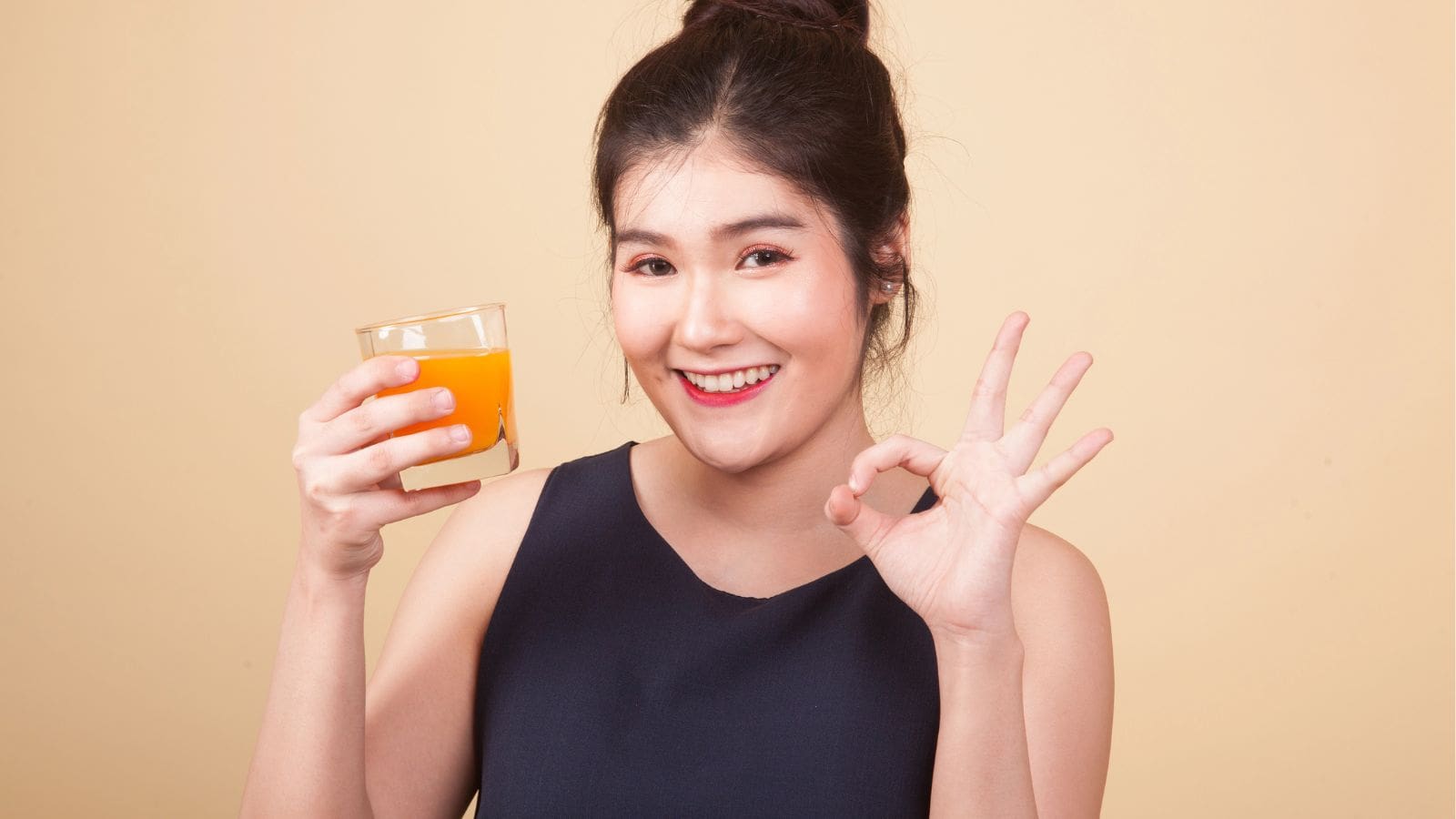 A woman with dark hair in a bun holds a glass of orange juice in one hand and makes an "OK" gesture with the other hand, smiling at the camera against a plain beige background.