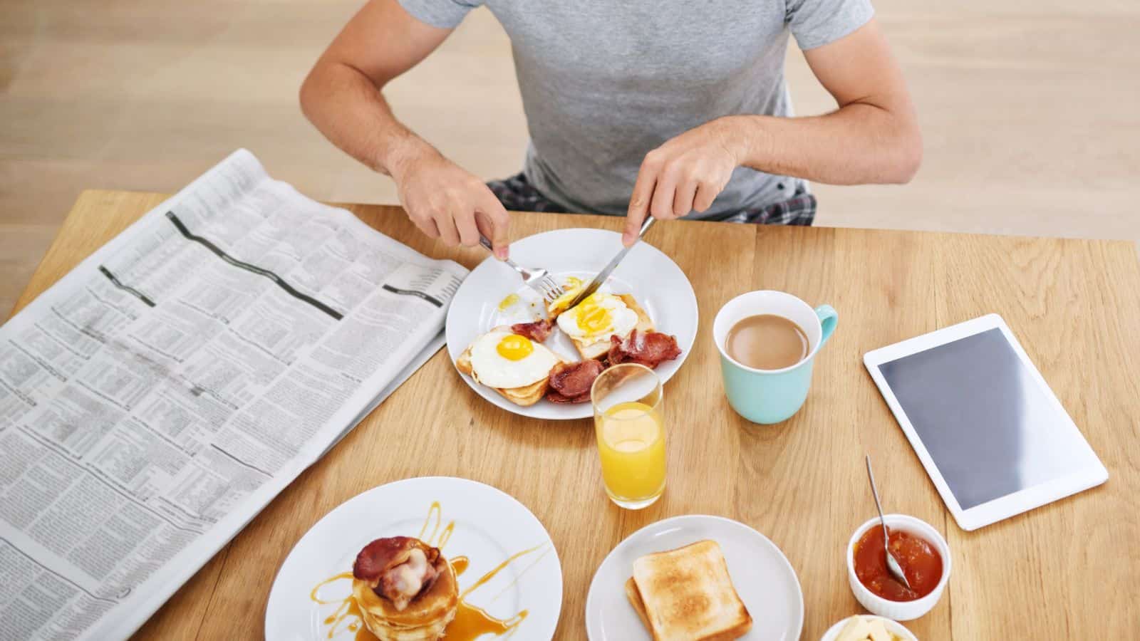A person sits at a wooden table eating eggs and bacon with toast, juice, and coffee. An open newspaper and a tablet are on the table along with pancakes and a small bowl of jam.