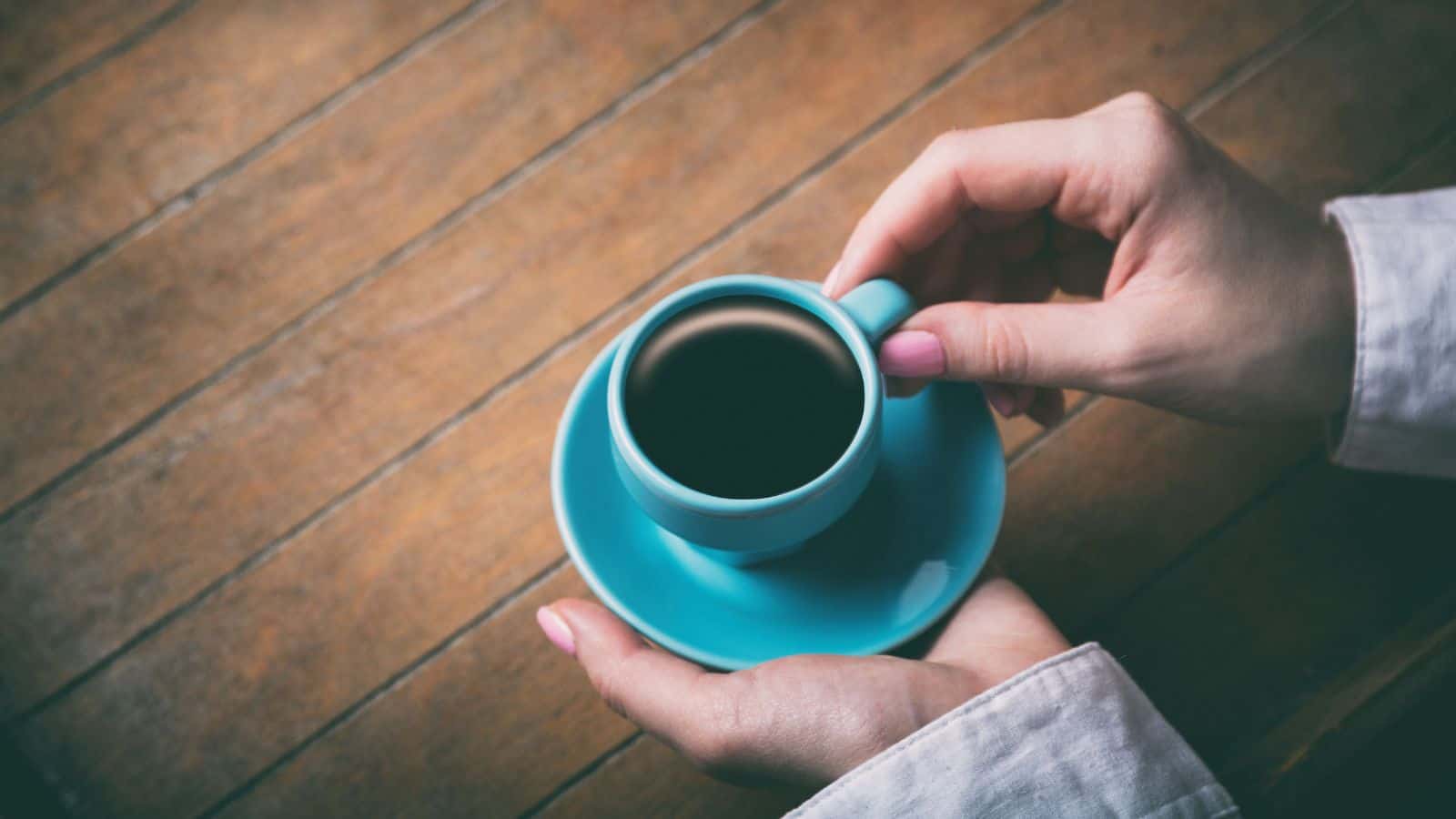 A person holding a small turquoise cup and saucer filled with coffee, resting their hands on a wooden table. The person is wearing a white long-sleeved shirt.