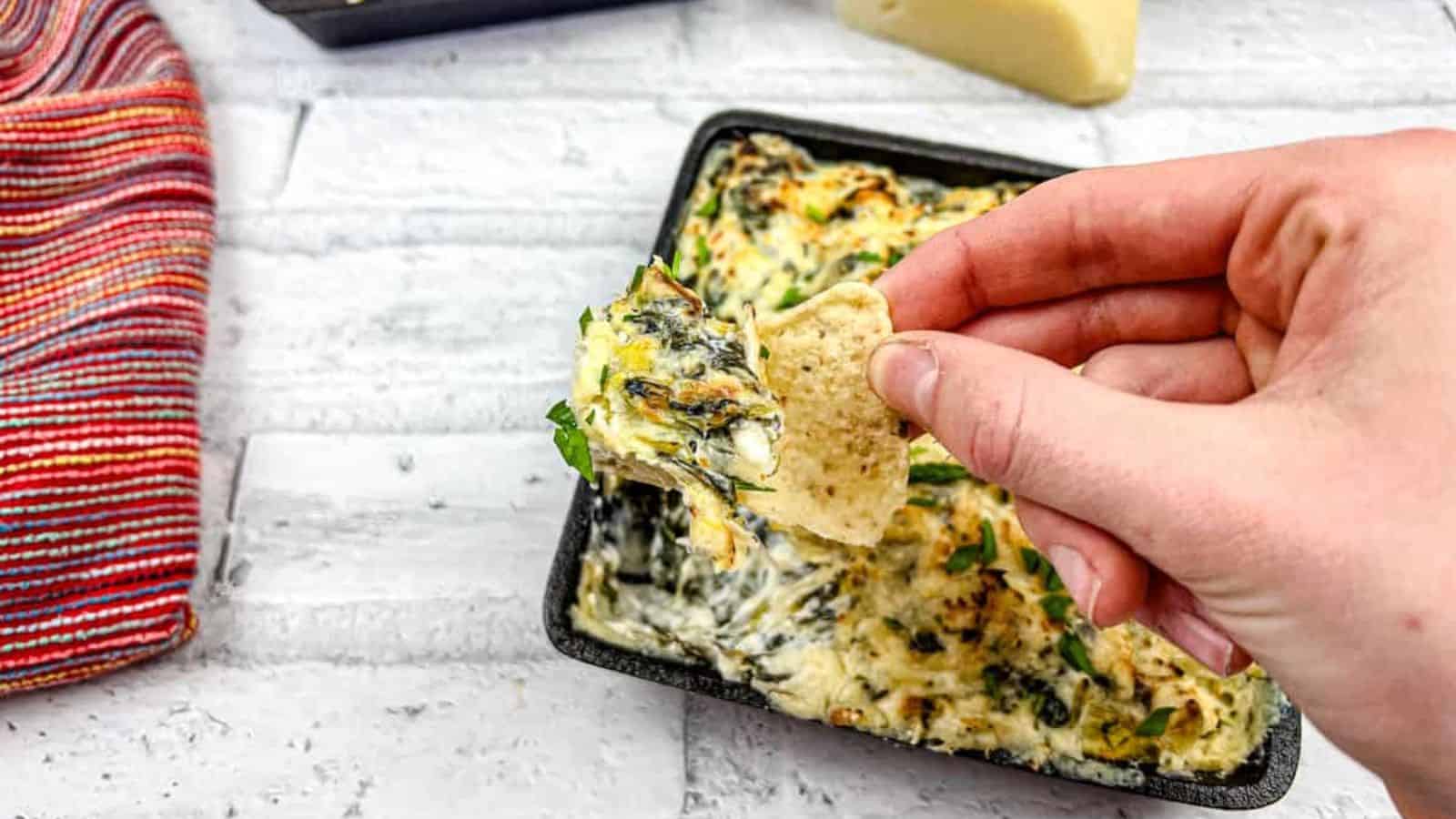 A hand dipping a tortilla chip into a tray of Copycat Applebee&rsquo;s spinach artichoke dip.