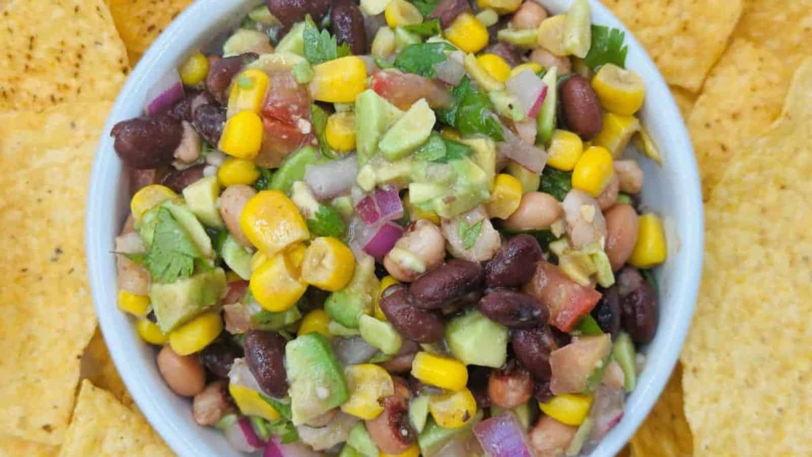 A bowl of bean and corn salad with diced avocado, red onion, tomatoes, and cilantro surrounded by tortilla chips.