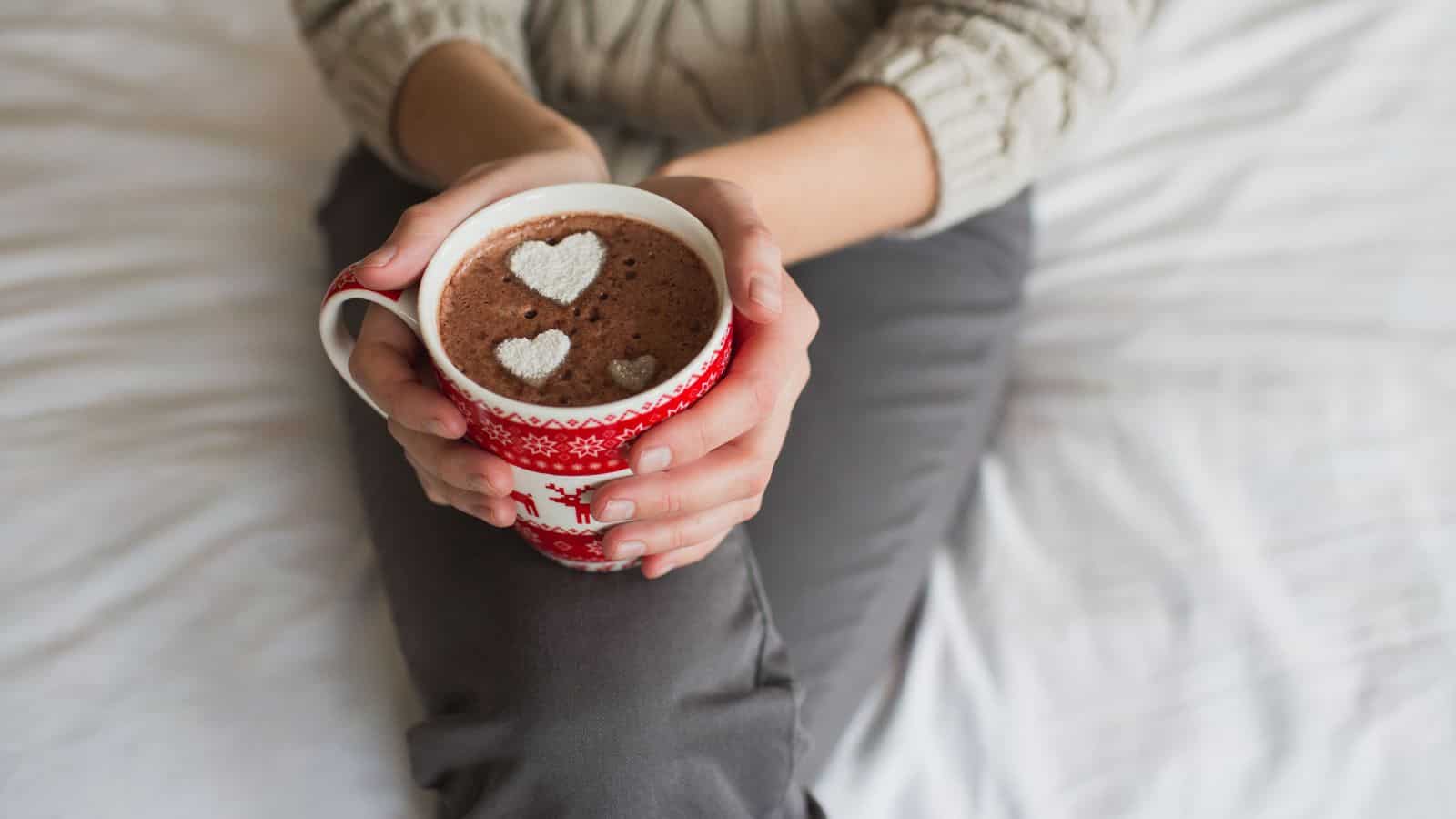 A person sitting on a bed holds a red mug with a white pattern, containing hot chocolate topped with foam in the shape of three hearts. The person is wearing a light sweater and gray pants.
