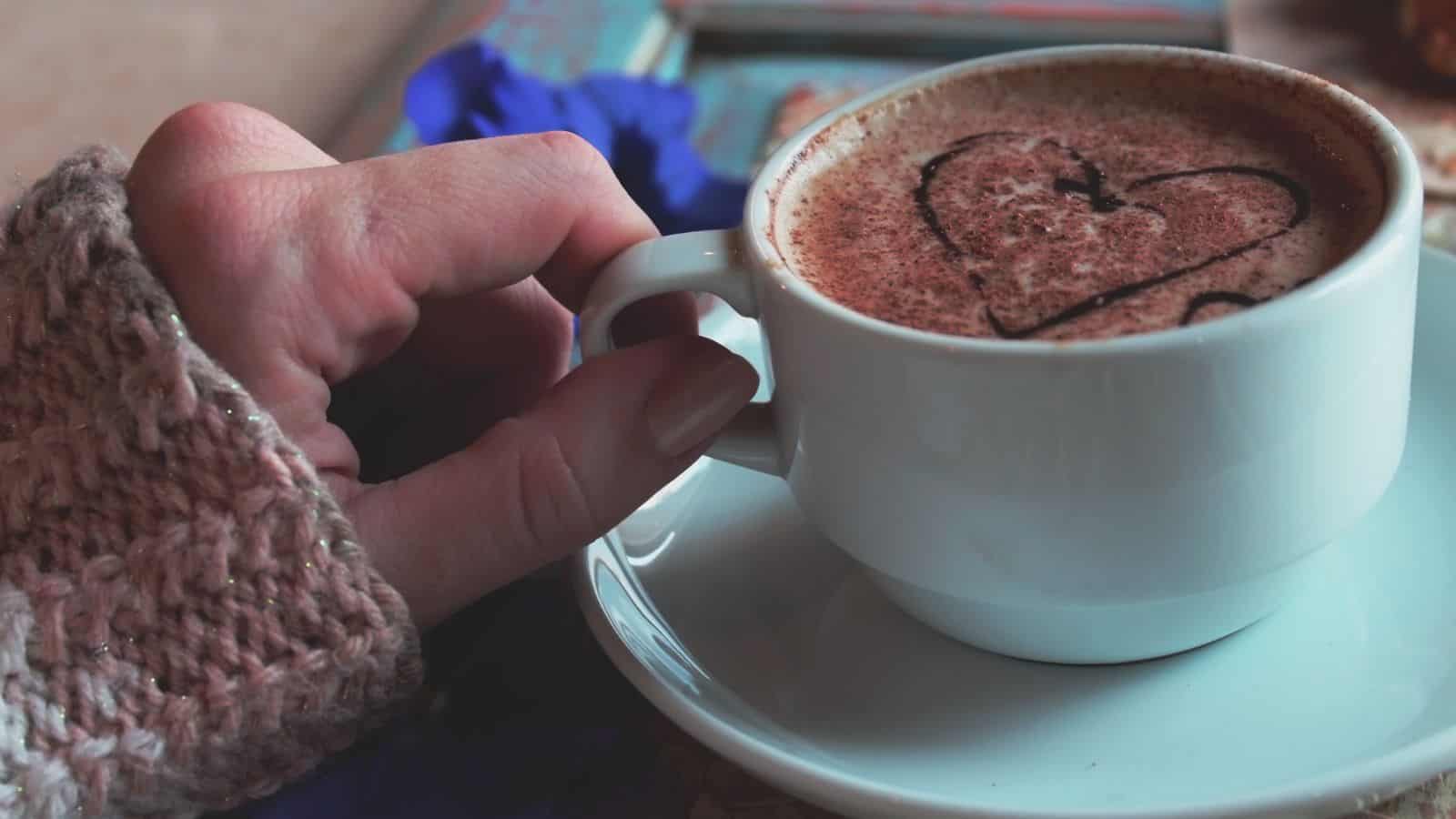 A hand in a knitted sleeve holds the handle of a white cup filled with frothy coffee, topped with a cocoa powder heart design. The cup rests on a white saucer, with a blurred blue flower in the background.