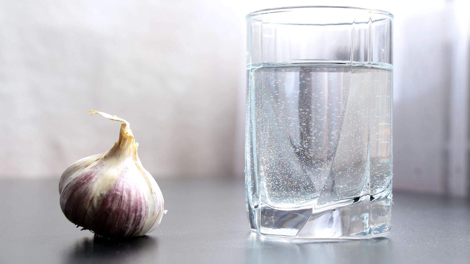 A whole garlic bulb sits on a smooth gray surface next to a clear glass of water. The glass is filled nearly to the top, with small bubbles visible in the water. The background is softly blurred.