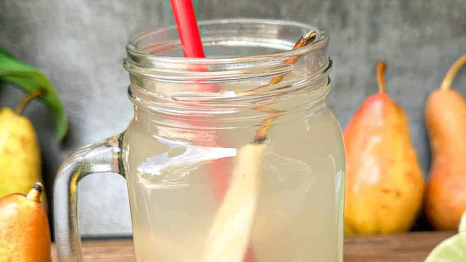 A glass mug filled with pear tea juice and a red straw, surrounded by whole pears, lime slices, and ginger on a wooden surface.