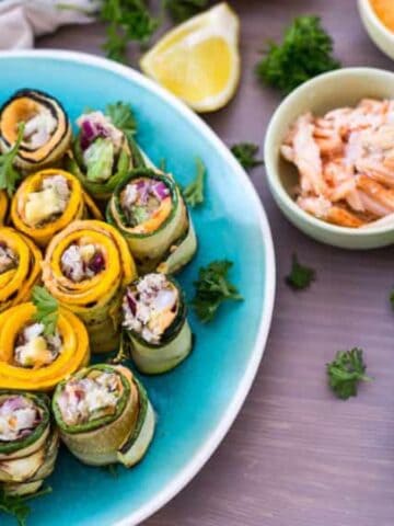 A blue plate with assorted vegetable roll-ups, garnished with herbs. Surrounding the plate are lemon wedges, fresh parsley, a bowl of shredded meat, a bowl of dip, and a bowl of chili flakes on a wooden surface.