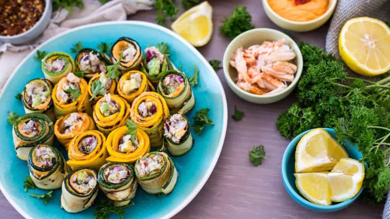 A blue plate with neatly arranged zucchini and yellow squash roll-ups filled with diced vegetables and herbs. Small bowls of ingredients and fresh parsley are visible in the background.