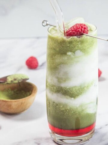 A tall glass with swirled layers of green and white iced drink, topped with two raspberries on a metal pick. Surrounding the glass are loose raspberries, a wooden spoon with green powder, a small bowl, and a bamboo whisk.