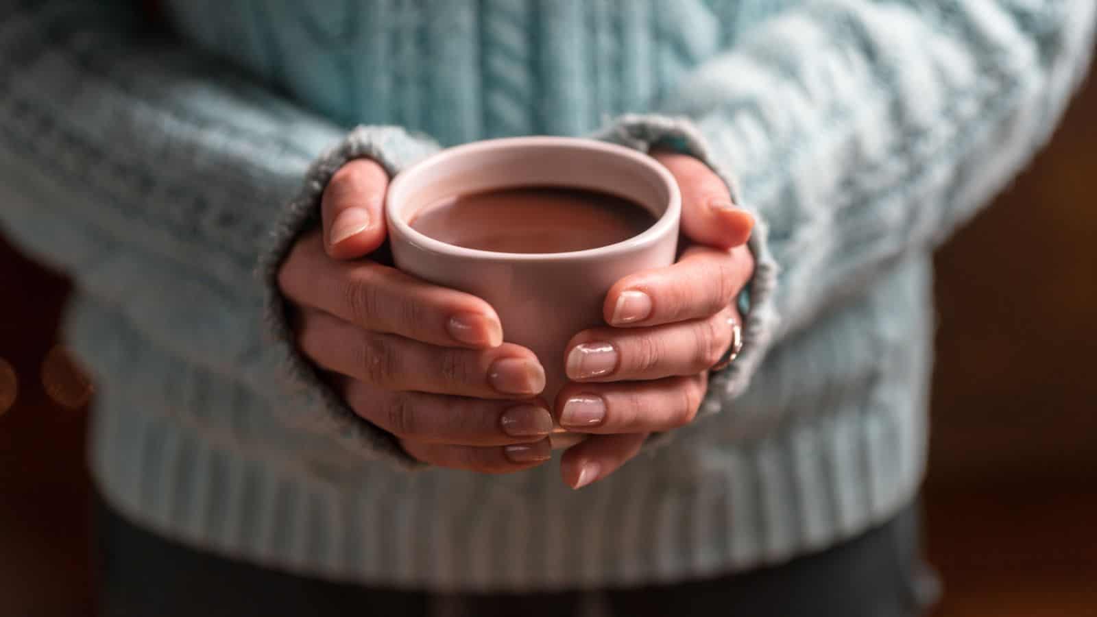 A person in a blue knitted sweater holds a white ceramic cup filled with a hot beverage, possibly cocoa or coffee, with both hands. The person's fingernails are neatly manicured.