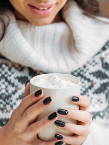 A person wearing a white and gray patterned sweater holds a white mug filled with marshmallows. The person has long dark hair and black nail polish, and is smiling slightly.