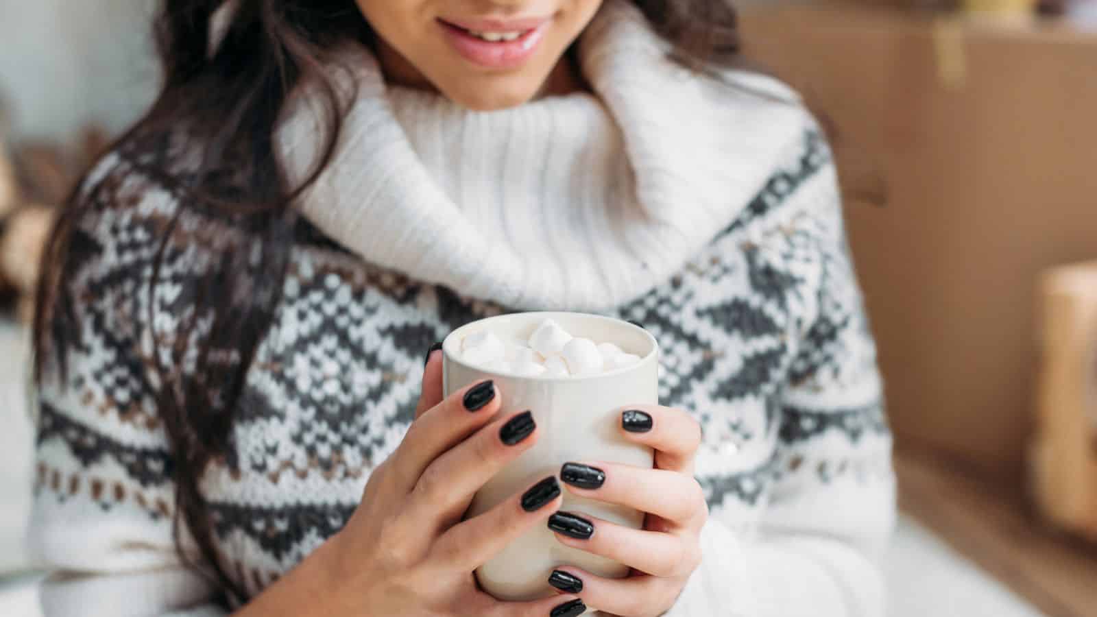A person wearing a white and gray patterned sweater holds a white mug filled with marshmallows. The person has long dark hair and black nail polish, and is smiling slightly.