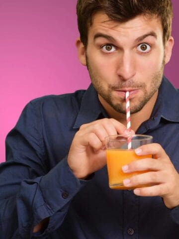 A man with brown hair wearing a blue shirt drinks orange juice from a glass using a red-and-white-striped straw, with a pink background behind him.