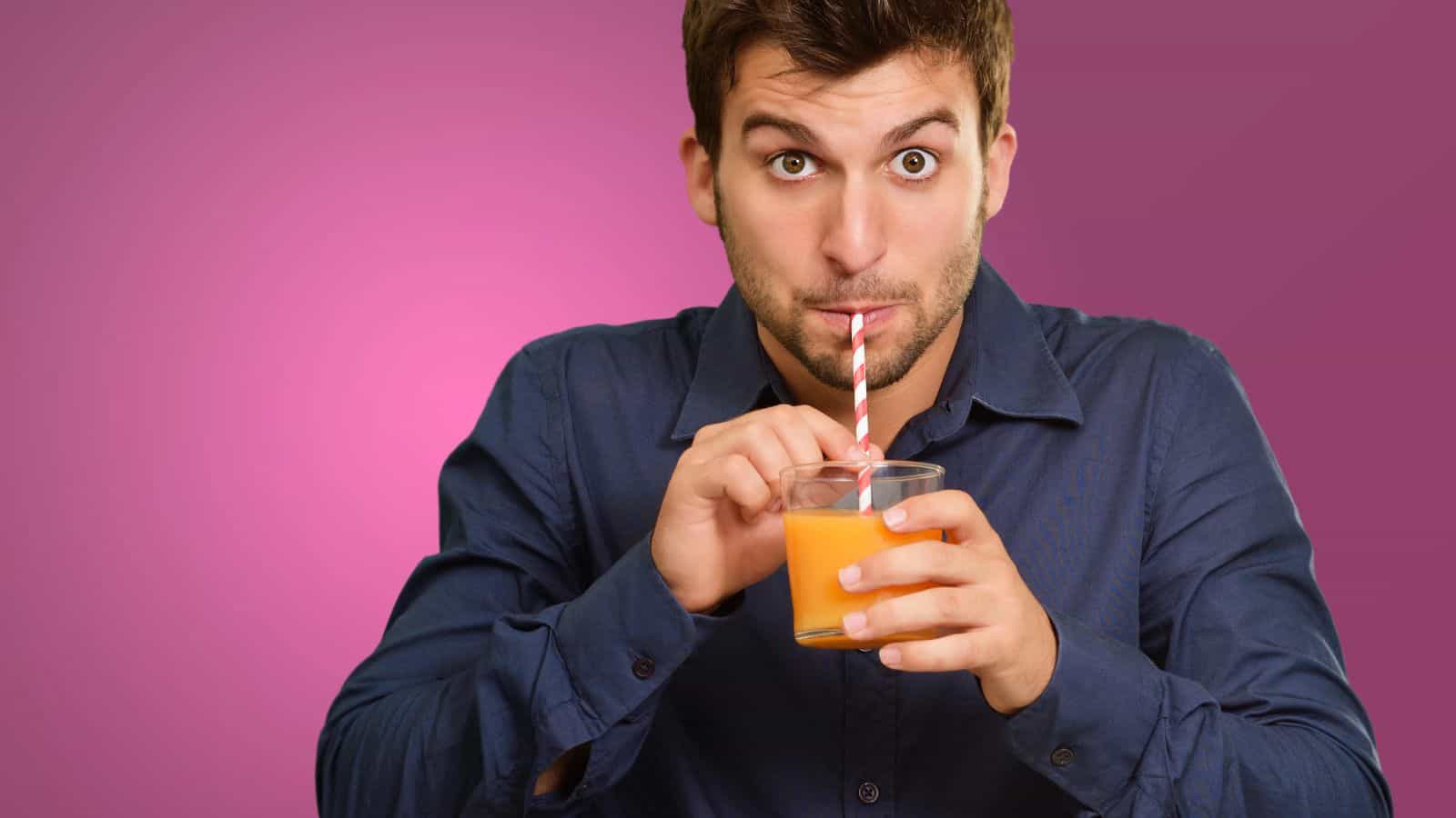 A man with brown hair wearing a blue shirt drinks orange juice from a glass using a red-and-white-striped straw, with a pink background behind him.