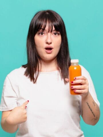 A person with straight dark hair and bangs, wearing a white shirt, holds an orange beverage in a clear plastic bottle and points at themselves, appearing surprised. The background is solid teal.