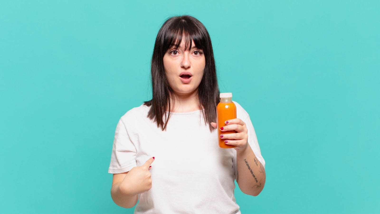 A person with straight dark hair and bangs, wearing a white shirt, holds an orange beverage in a clear plastic bottle and points at themselves, appearing surprised. The background is solid teal.