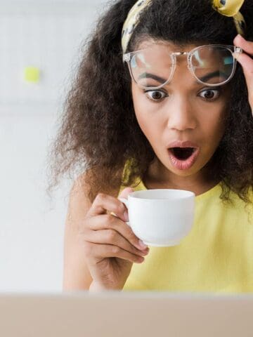 A woman with curly hair raises her glasses in surprise while holding a white teacup and looking at a laptop screen. She is wearing a yellow top and a white watch, sitting in a bright room.