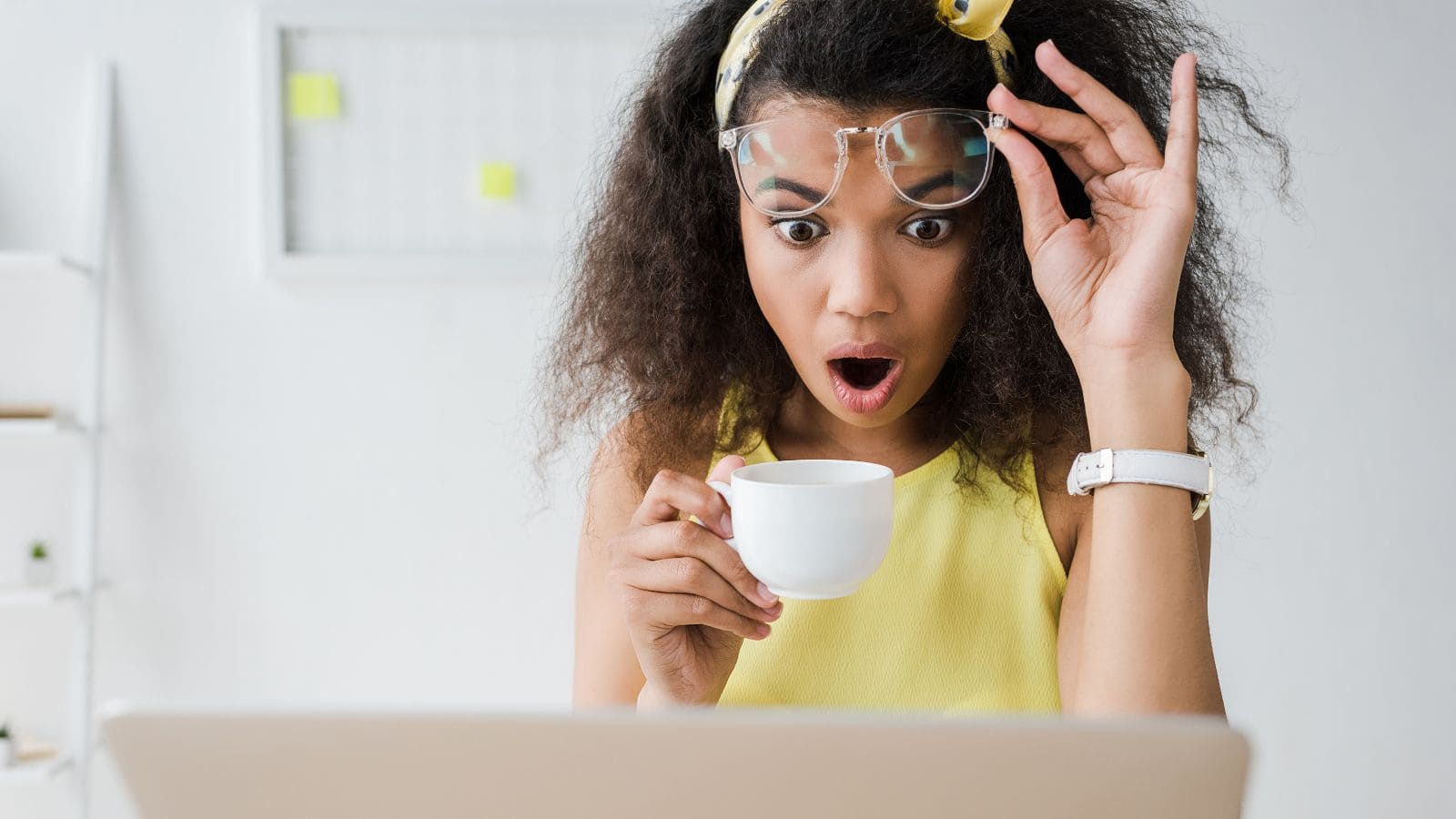 A woman with curly hair raises her glasses in surprise while holding a white teacup and looking at a laptop screen. She is wearing a yellow top and a white watch, sitting in a bright room.