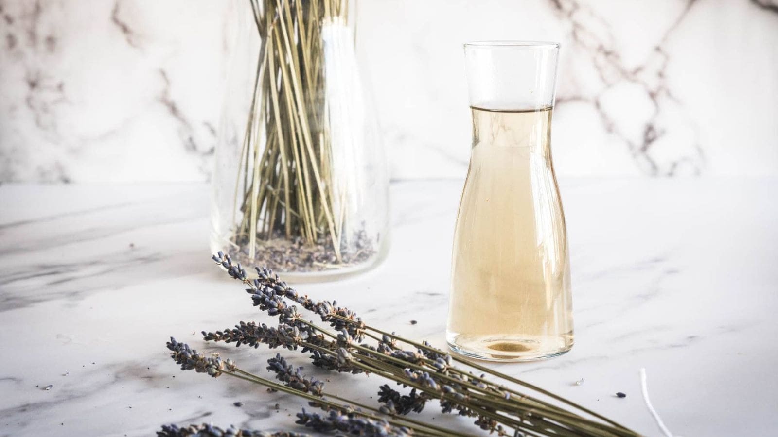 A clear glass carafe filled with light-colored liquid sits on a white marble surface. Nearby are dried lavender sprigs and a glass vase holding more dried lavender stems.