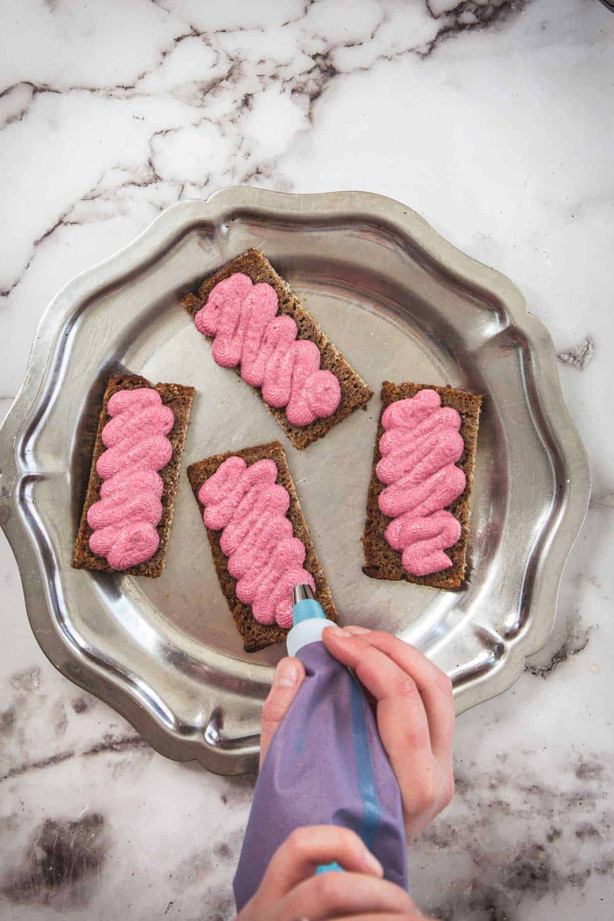 A person pipes pink frosting in a zigzag pattern onto four rectangular pieces of crispbread arranged on a silver, scalloped-edged plate set on a marble surface.
