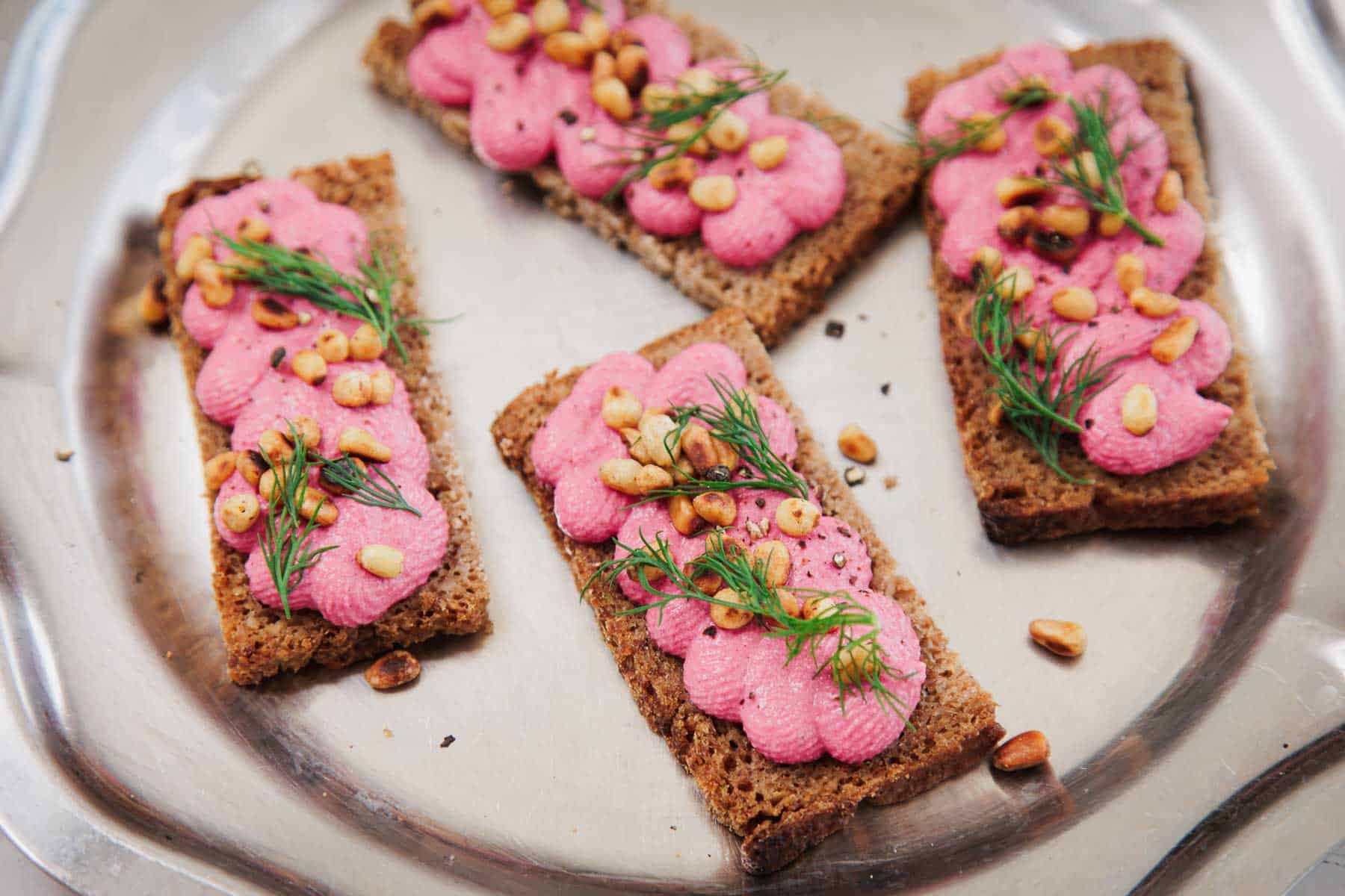 Four pieces of rectangular rye bread are topped with a pink spread, pine nuts, and fresh dill, arranged on a round metal tray.