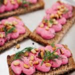 Rectangular pieces of dark crispbread are topped with a bright pink spread, garnished with pine nuts, black pepper, and fresh dill, and arranged on a metal tray.