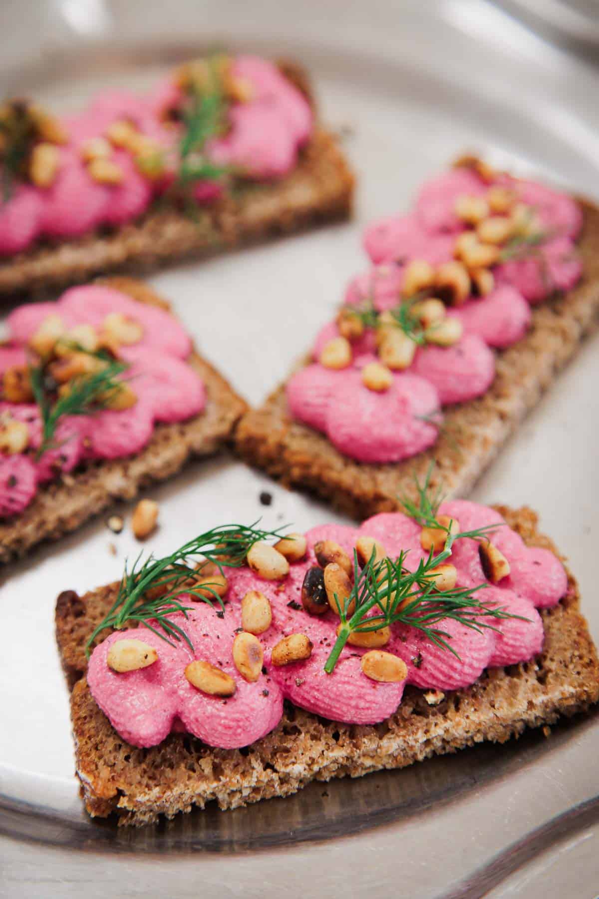 Rectangular pieces of dark crispbread are topped with a bright pink spread, garnished with pine nuts, black pepper, and fresh dill, and arranged on a metal tray.