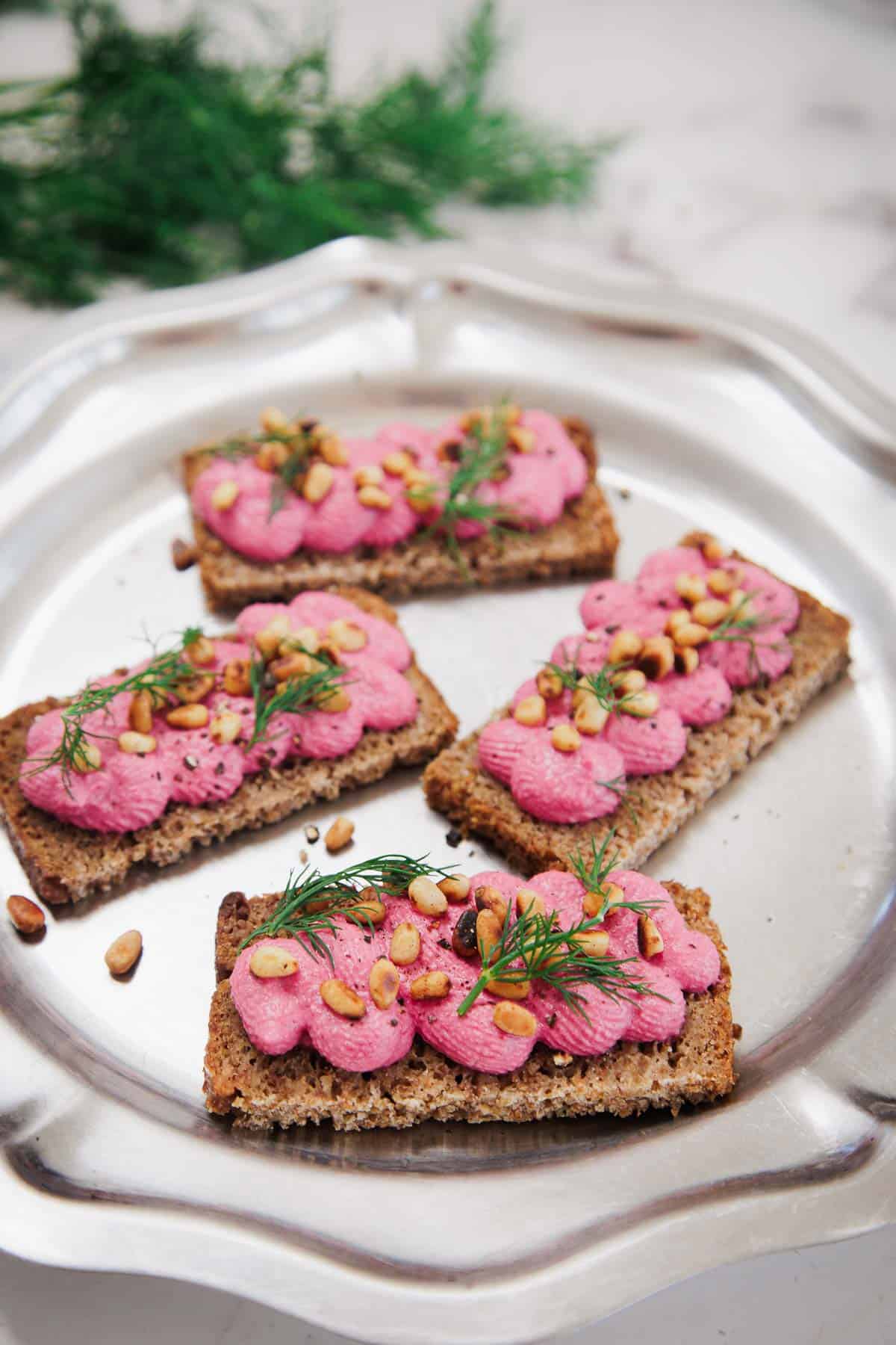 Four rectangular slices of dark bread topped with bright pink spread, pine nuts, and dill sprigs are arranged on a silver plate. A green herb is blurred in the background.