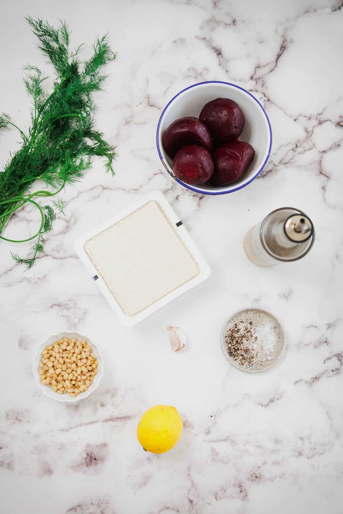 A block of tofu, a bowl of whole beets, fresh dill, a small bowl of pine nuts, a lemon, a clove of garlic, salt and pepper in a dish, and a bottle of olive oil arranged on a white marble countertop.