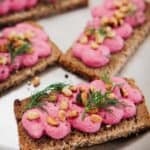 Rectangular pieces of dark bread topped with bright pink spread, pine nuts, fresh dill, and ground black pepper are arranged on a metal tray. The spread is piped in a decorative pattern on each piece.
