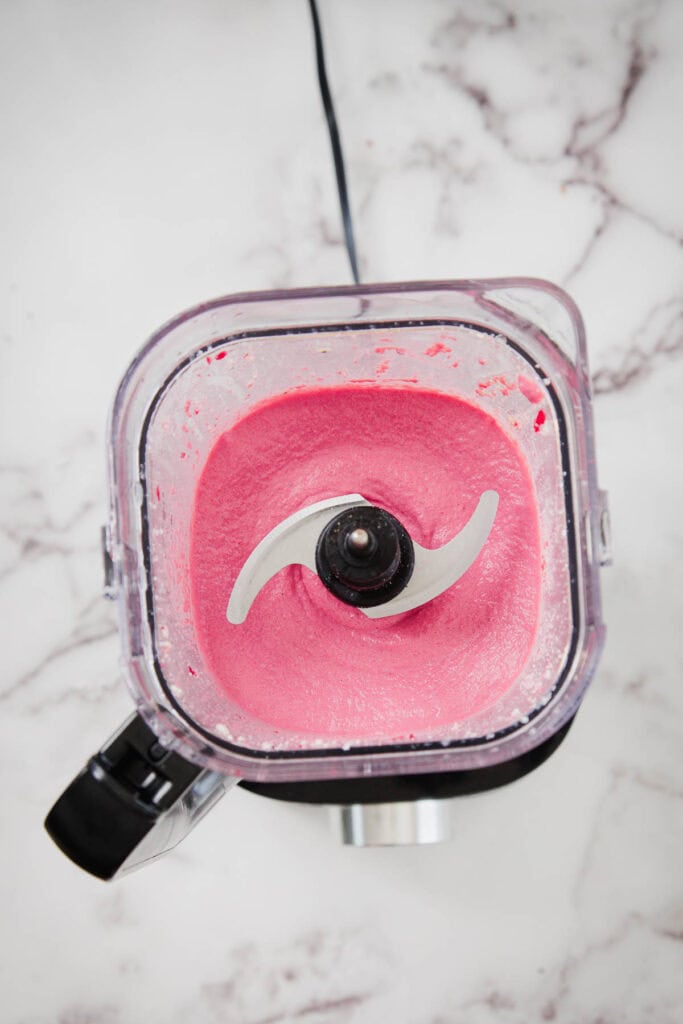 A top-down view of a blender on a marble surface, containing a smooth, thick, bright pink mixture. The blender&rsquo;s sharp blade is visible in the center.