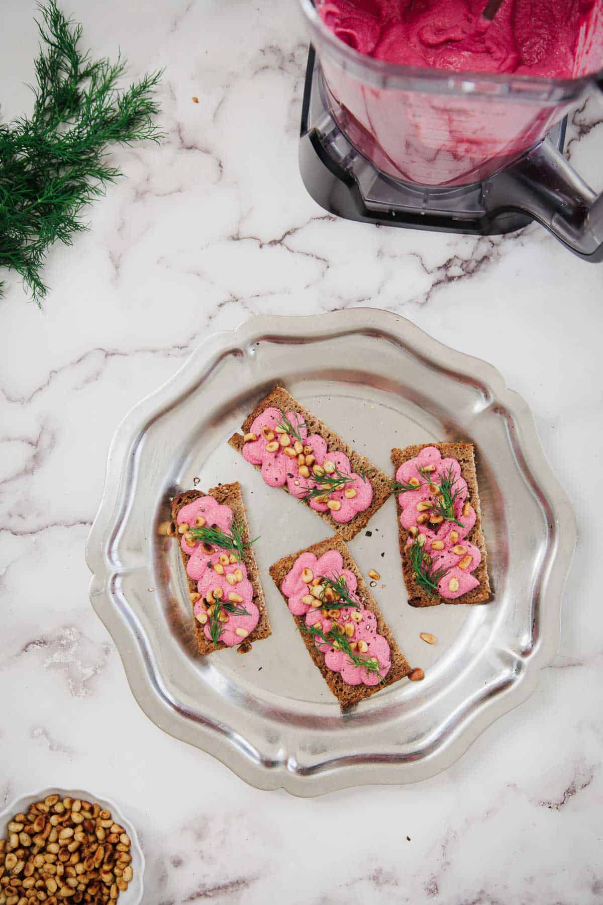 Four rectangular crackers topped with a pink spread, fresh dill, and pine nuts are arranged on a silver plate. A food processor with leftover pink spread and a bowl of pine nuts are nearby on a marble surface.