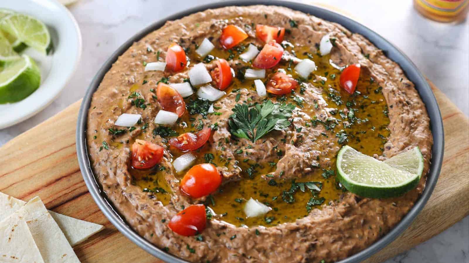A bowl of refried beans topped with chopped tomatoes, diced onions, fresh parsley, and a lime wedge sits on a wooden board. Tortilla pieces and lime slices are visible nearby.
