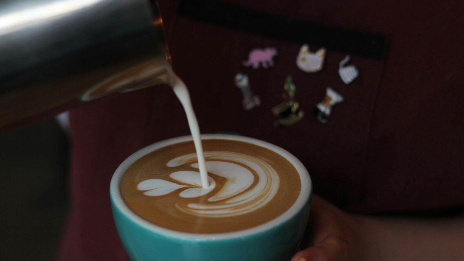 A close-up of milk being poured into a cup of coffee with latte art forming on top; the person pouring wears a dark apron with several animal-shaped pins on it.