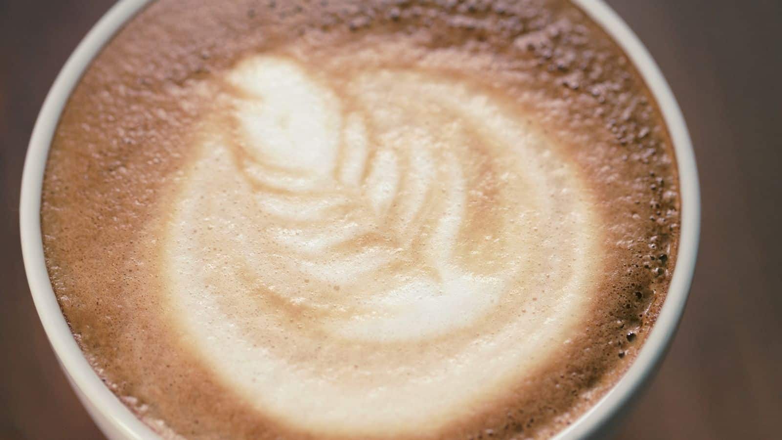 A close-up view of a cup of cappuccino with a latte art design in the foam, showing a light brown and creamy white pattern on the surface.