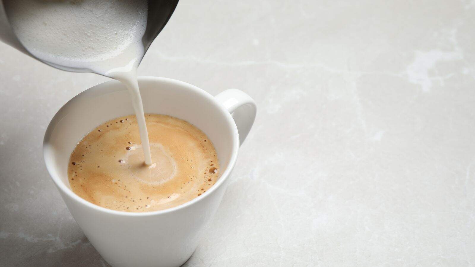 A close-up of steamed milk being poured from a metal pitcher into a white cup of coffee on a light, textured surface.