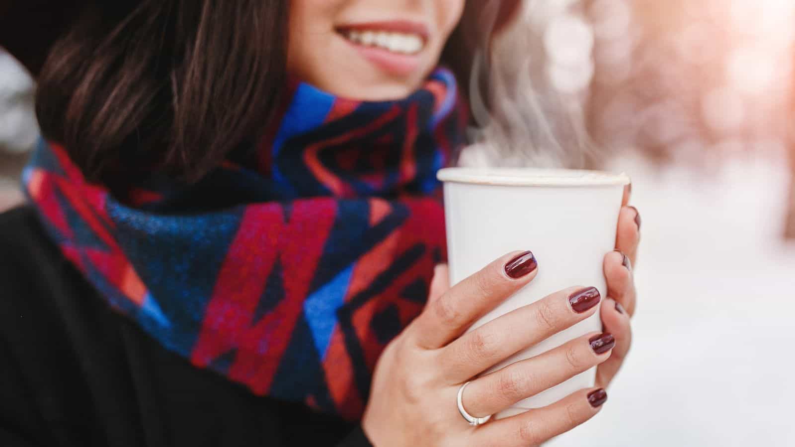 A person with dark hair and painted nails holds a steaming white cup with both hands. They are wearing a colorful scarf and dark clothing, and they are smiling. The background appears to be outdoors in winter.