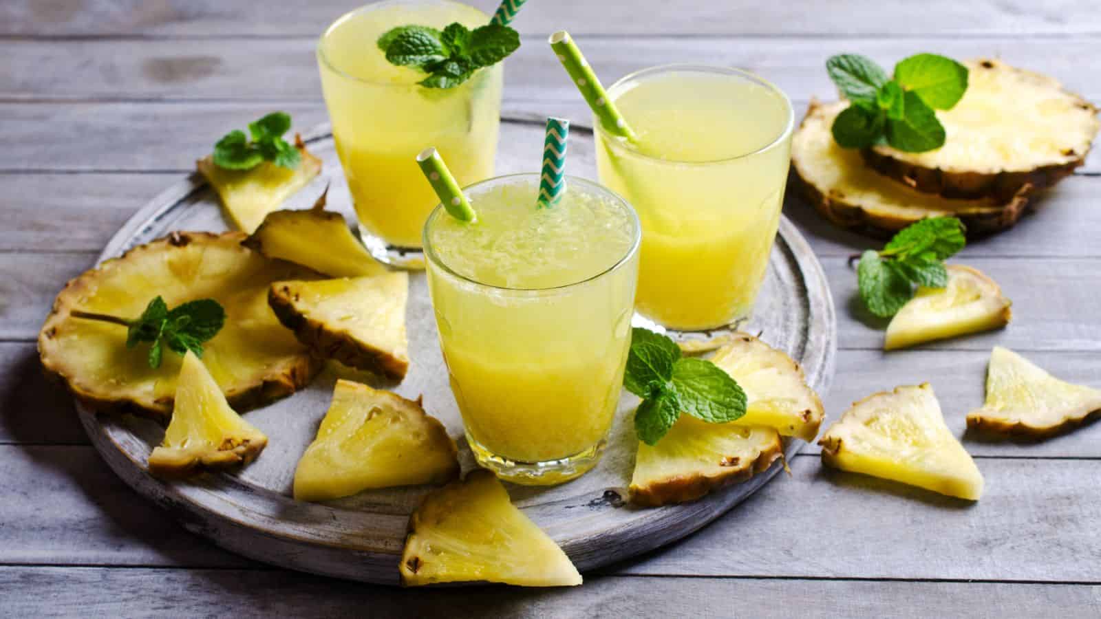 Three glasses of pineapple juice with straws and mint leaves on top, placed on a round tray with several fresh pineapple slices and mint sprigs, set on a wooden surface.