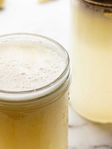 Two glass jars filled with a pale yellow, frothy liquid are placed on a white marble surface. Lemon wedges are scattered in the background.