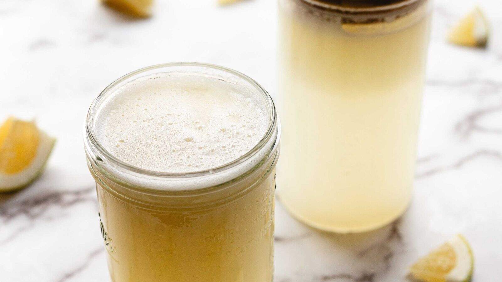 Two glass jars filled with a pale yellow, frothy liquid are placed on a white marble surface. Lemon wedges are scattered in the background.